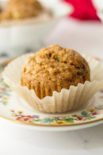 A single muffin in a paper liner sits on a floral-patterned plate, with more muffins and a teacup blurred in the background, creating a cozy scene perfect for enjoying freshly baked muffins.