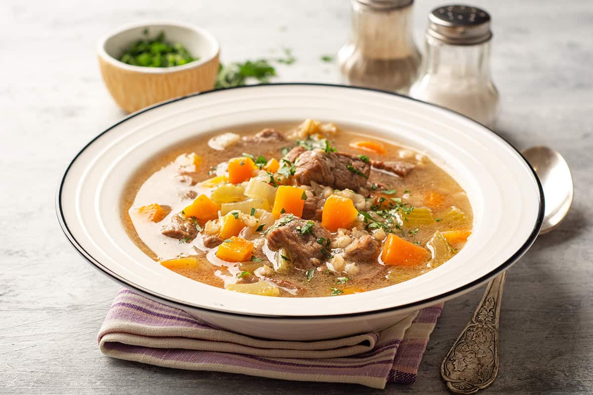 A bowl of beef barley and vegetable stew with carrots and herbs, placed on a striped napkin, with a spoon and seasonings in the background.