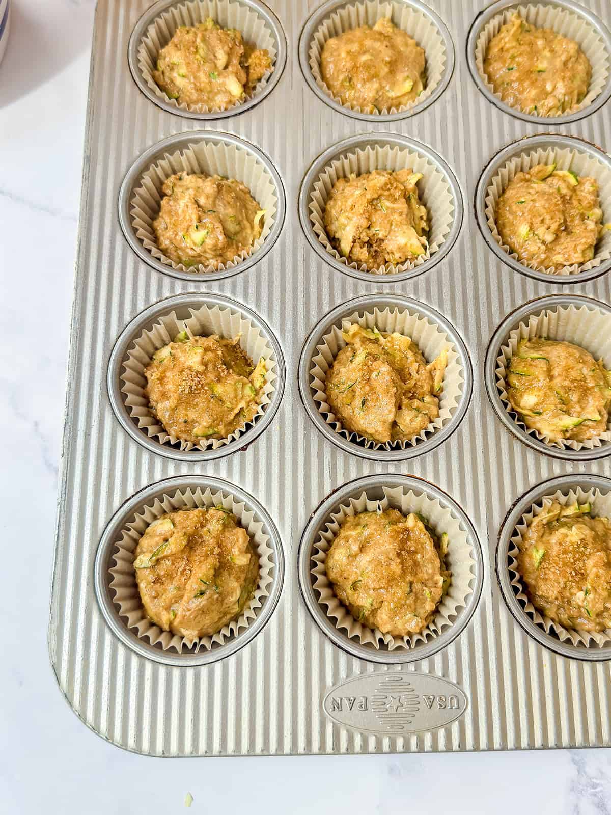Muffin tin with paper liners filled with unbaked muffins batter, placed on a white countertop.