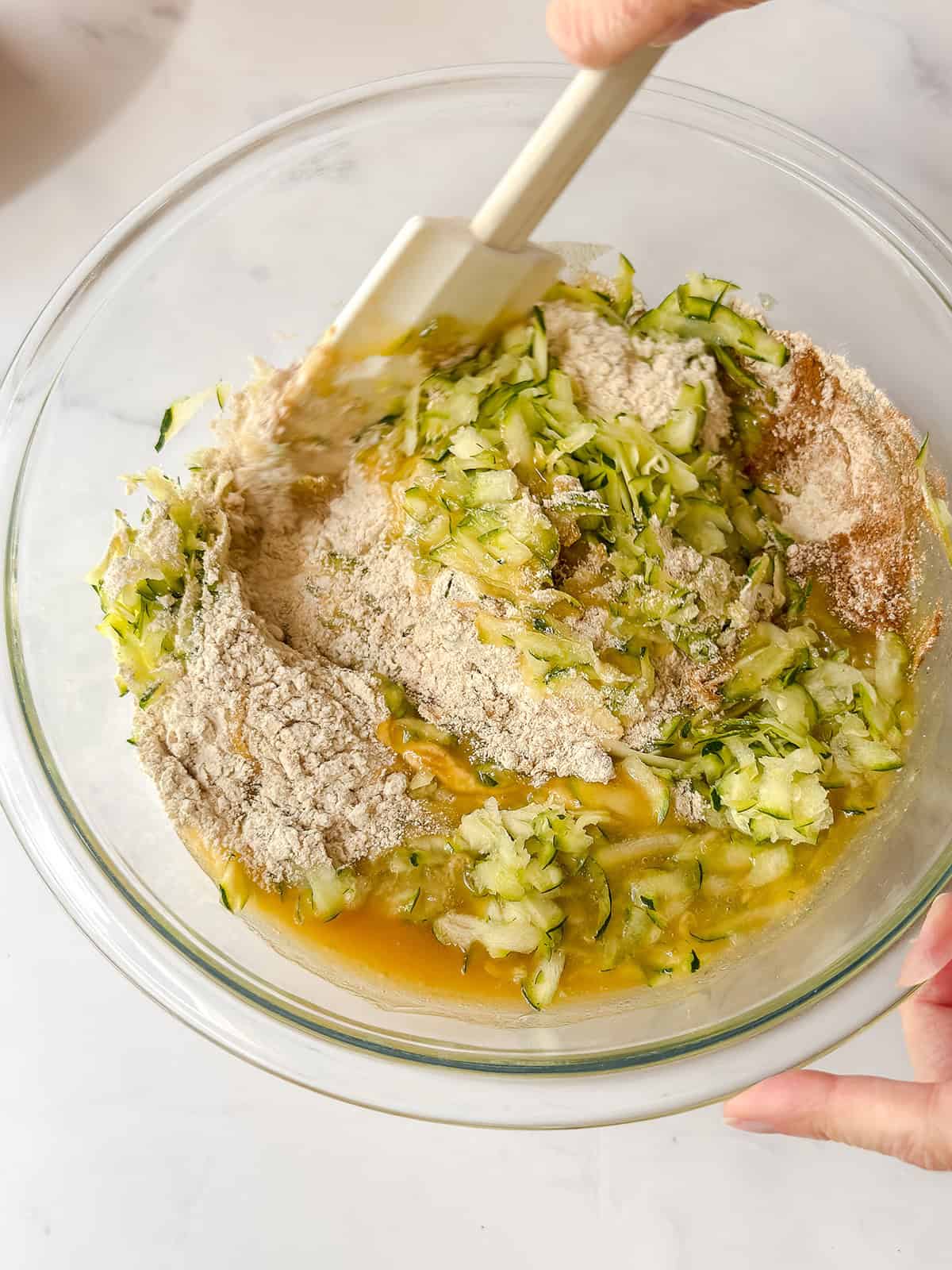 A hand uses a spatula to mix shredded zucchini, flour, wet ingredients, and muffin batter in a glass bowl on a white surface.