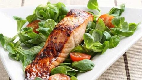 A cooked Wild King Salmon fillet rests on a bed of green leafy salad with halved cherry tomatoes, served on a white rectangular plate.