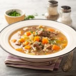A bowl of beef barley and vegetable soup with diced carrots and herbs, placed on a striped napkin, with salt, pepper shakers, and a small bowl of chopped herbs in the background.