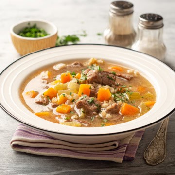 A bowl of beef barley and vegetable soup with diced carrots and herbs, placed on a striped napkin, with salt, pepper shakers, and a small bowl of chopped herbs in the background.