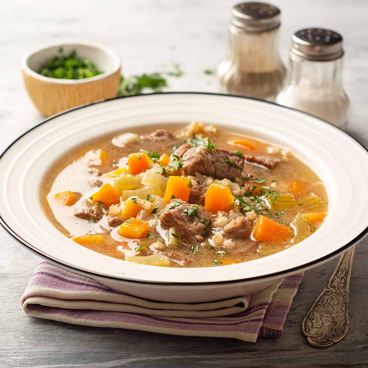 A bowl of beef barley and vegetable soup with diced carrots and herbs, placed on a striped napkin, with salt, pepper shakers, and a small bowl of chopped herbs in the background.