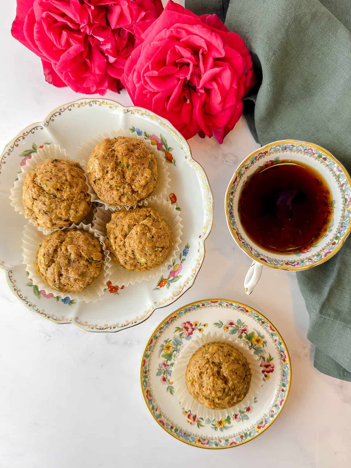 A floral plate with five golden muffins sits next to a teacup of dark tea, while a matching saucer holds one more muffin. 