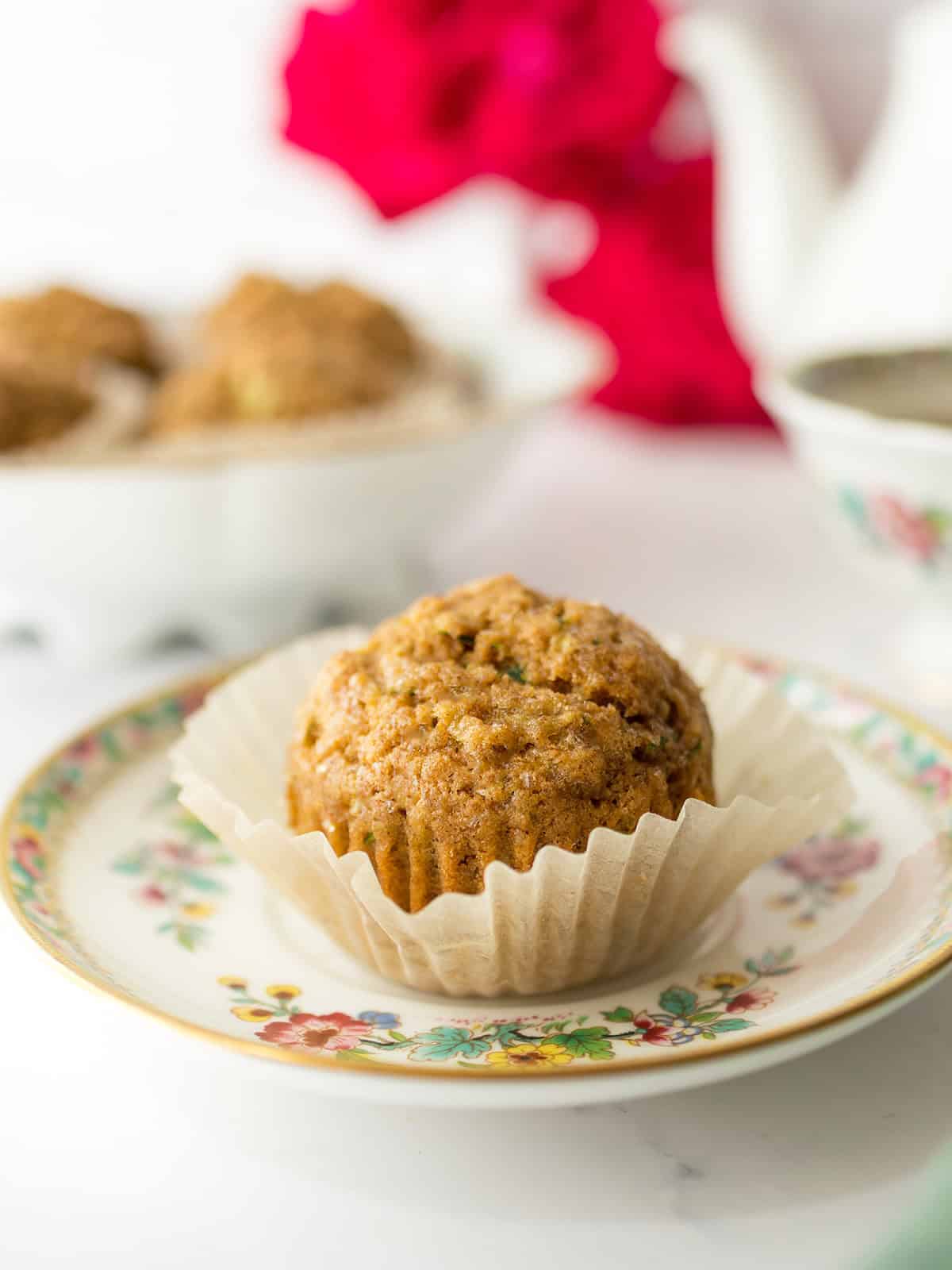 A single zucchini muffin in a paper liner sits on a floral plate, with more freshly baked muffins and a teapot blurred in the background.