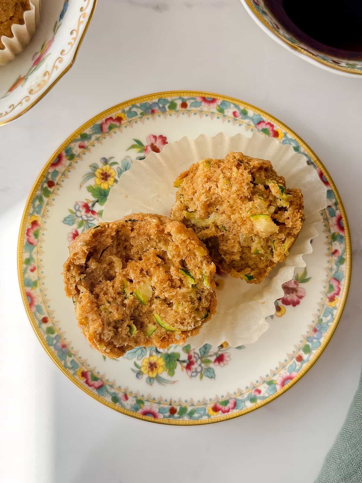 Zucchini muffin split in half on a floral patterned plate with a cupcake liner, next to a cup of tea.