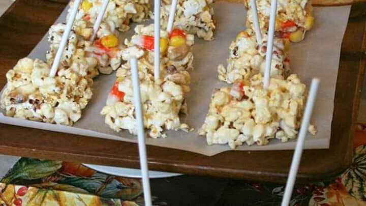 Halloween popcorn treats with colorful candy corn on sticks are arranged on a tray, set on an autumn-themed tablecloth. Pumpkins and gourds loom in the background, enhancing the festive seasonal feel.