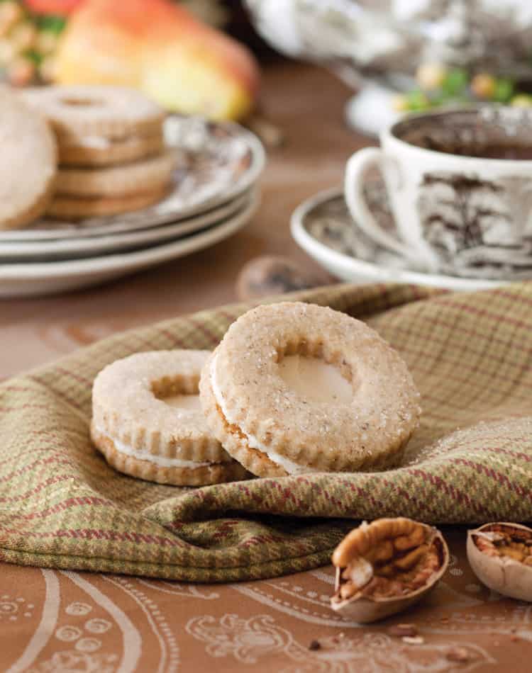 Two round sandwich cookies dusted with sugar rest on a plaid cloth, perfect for an Autumn Afternoon Tea. A cup of coffee and plates of cookies sit in the background, while broken nuts are scattered in the foreground.