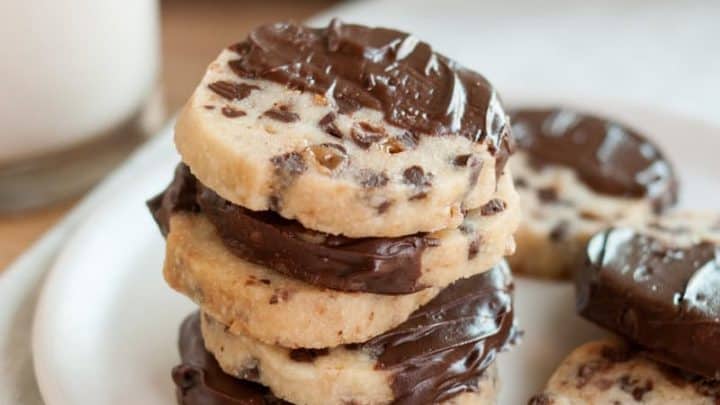 A stack of chocolate chip cookies half-dipped in chocolate sits on a white plate next to a glass of milk—perfect Make Ahead Christmas Cookies. More cookies are visible in the background.