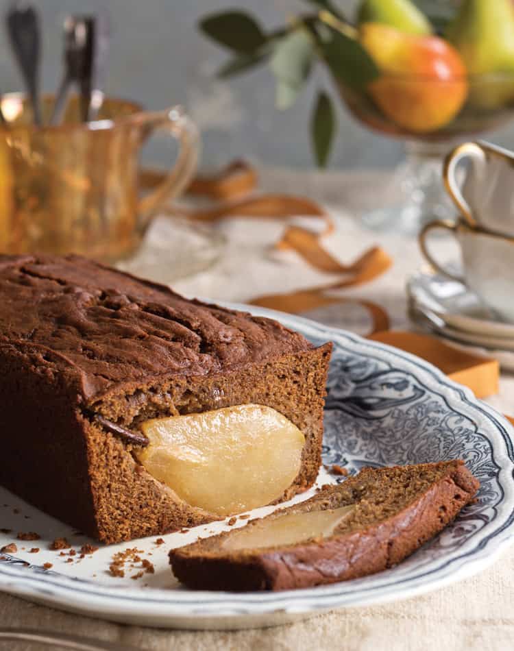A loaf of cake with a whole pear baked inside, served on a patterned plate with a sliced piece in front-perfect for an Autumn Afternoon Tea with teacups and fruit elegantly arranged in the background.