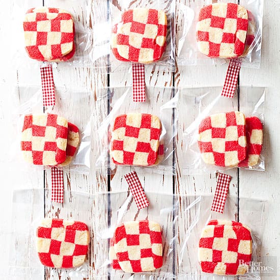 Checkerboard cookies in festive red and cream, perfect as make-ahead Christmas treats, are individually wrapped in clear bags with red gingham ribbon ties and arranged in a grid pattern on a white wooden surface.