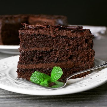 A slice of rich chocolate cake layered with chocolate shavings and a mint garnish sits on a white plate with a fork.