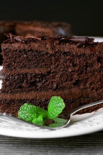 A slice of rich chocolate cake layered with chocolate shavings and a mint garnish sits on a white plate with a fork.