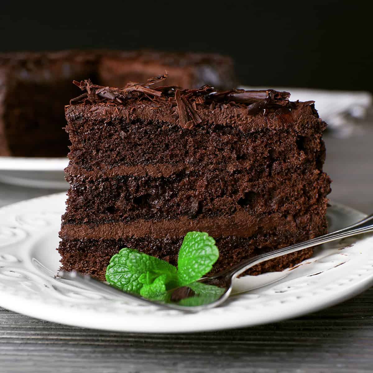 A slice of rich chocolate cake layered with chocolate shavings and a mint garnish sits on a white plate with a fork.