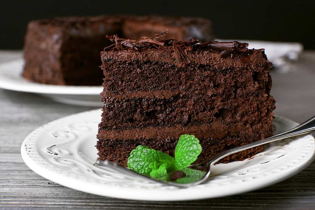 A slice of decadent chocolate cake with rich frosting and chocolate shavings sits on a white plate, garnished with mint leaves and served with a fork.