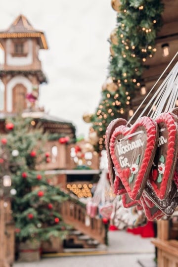 Square image of European Christmas market featuring Christmas Market foods with a bell tower in the background and a decorated Christmas tree.