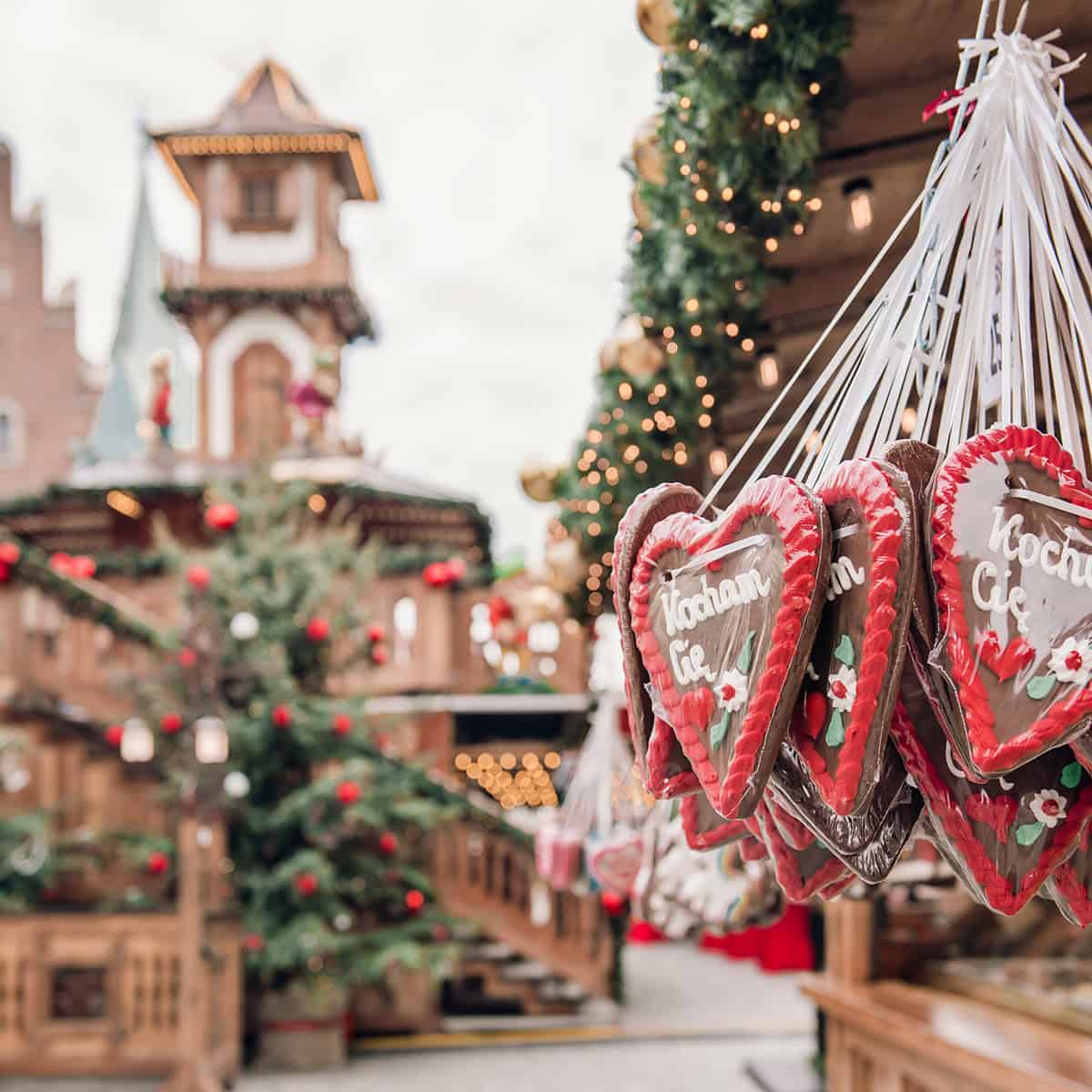 Square image of European Christmas market featuring Christmas Market foods with a bell tower in the background and a decorated Christmas tree.