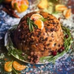 Traditional English Christmas Pudding on a serving dish with fresh rosemary, dried fruit, and orange segments as garnish for serving.