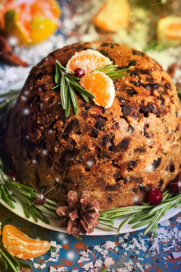 Traditional English Christmas Pudding on a serving dish with fresh rosemary, dried fruit, and orange segments as garnish for serving.