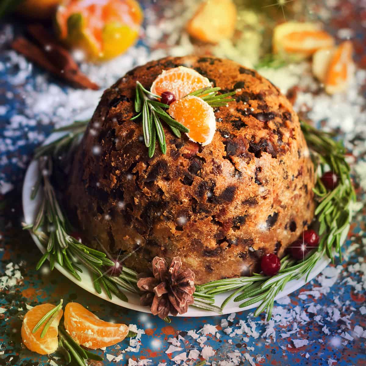 Traditional English Christmas Pudding on a serving dish with fresh rosemary, dried fruit, and orange segments as garnish for serving.