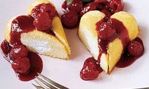 A heart-shaped sponge cake with white filling, topped with cherry sauce and garnished with mint, is served on a white plate with two forks—a perfect addition to your Valentine's Day treats.