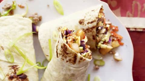 Two grilled wraps, each crafted with just 5 ingredients, rest on a white plate filled with colorful vegetables and garnished with sliced green onions. A small bowl of dark sauce peeks from the background, all elegantly set atop a patterned red tablecloth.