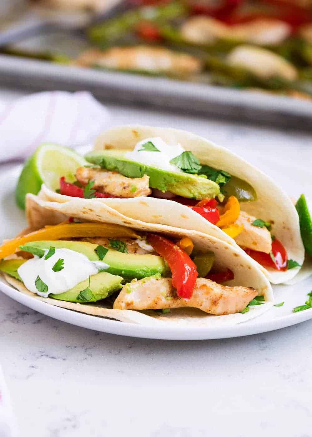 Two soft flour tortillas filled with 5 ingredient grilled chicken strips, sliced avocado, bell peppers, fresh cilantro, and a dollop of sour cream, served on a white plate.