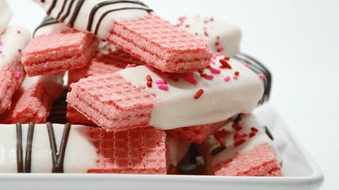 A stack of pink wafer cookies, partially dipped in white chocolate and adorned with chocolate drizzle and colorful sprinkles, arranged on a white rectangular plate—perfect as festive Valentine's Day treats.