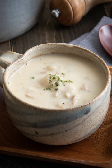 A ceramic mug filled with creamy fish chowder garnished with herbs, placed on a wooden plate with a spoon and napkin nearby.