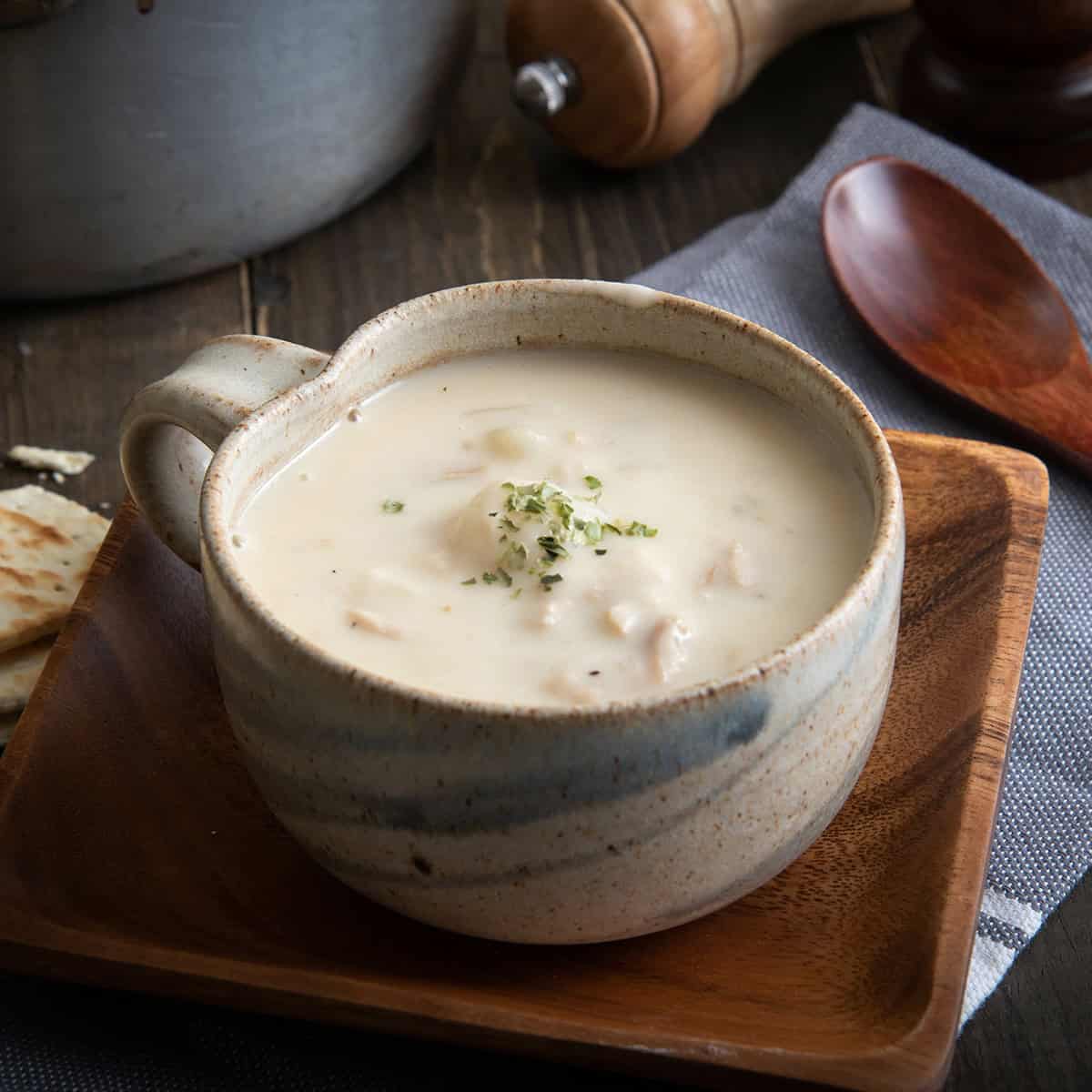 A ceramic mug filled with creamy fish chowder garnished with herbs, placed on a wooden plate with a spoon and napkin nearby.