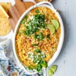 An oval dish of cheesy corn dip—perfect for Game Day Dips—topped with cilantro, crumbled cheese, chili powder, and lime wedges on a white wooden surface.