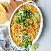 An oval dish of cheesy corn dip-perfect for Game Day Dips-topped with cilantro, crumbled cheese, chili powder, and lime wedges on a white wooden surface.