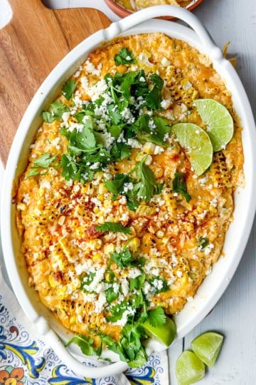 An oval dish of cheesy corn dip—perfect for Game Day Dips—topped with cilantro, crumbled cheese, chili powder, and lime wedges on a white wooden surface.