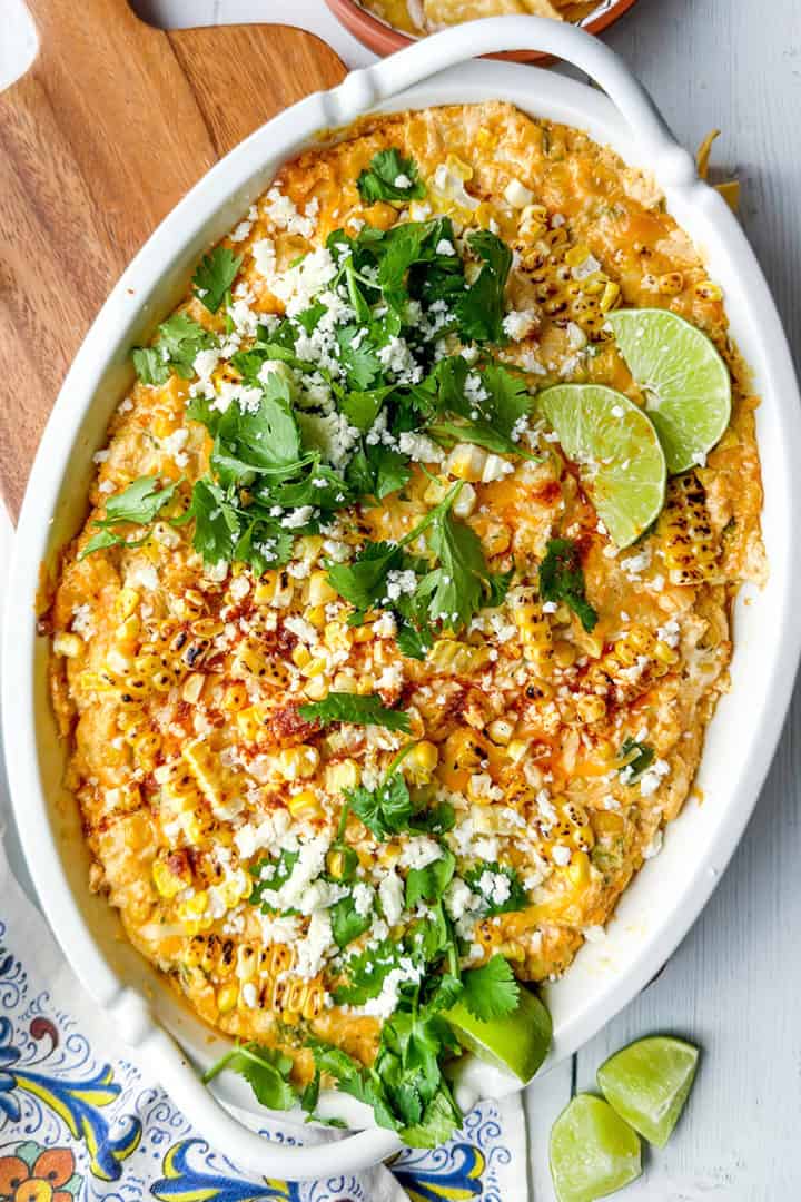 An oval dish of cheesy corn dip—perfect for Game Day Dips—topped with cilantro, crumbled cheese, chili powder, and lime wedges on a white wooden surface.