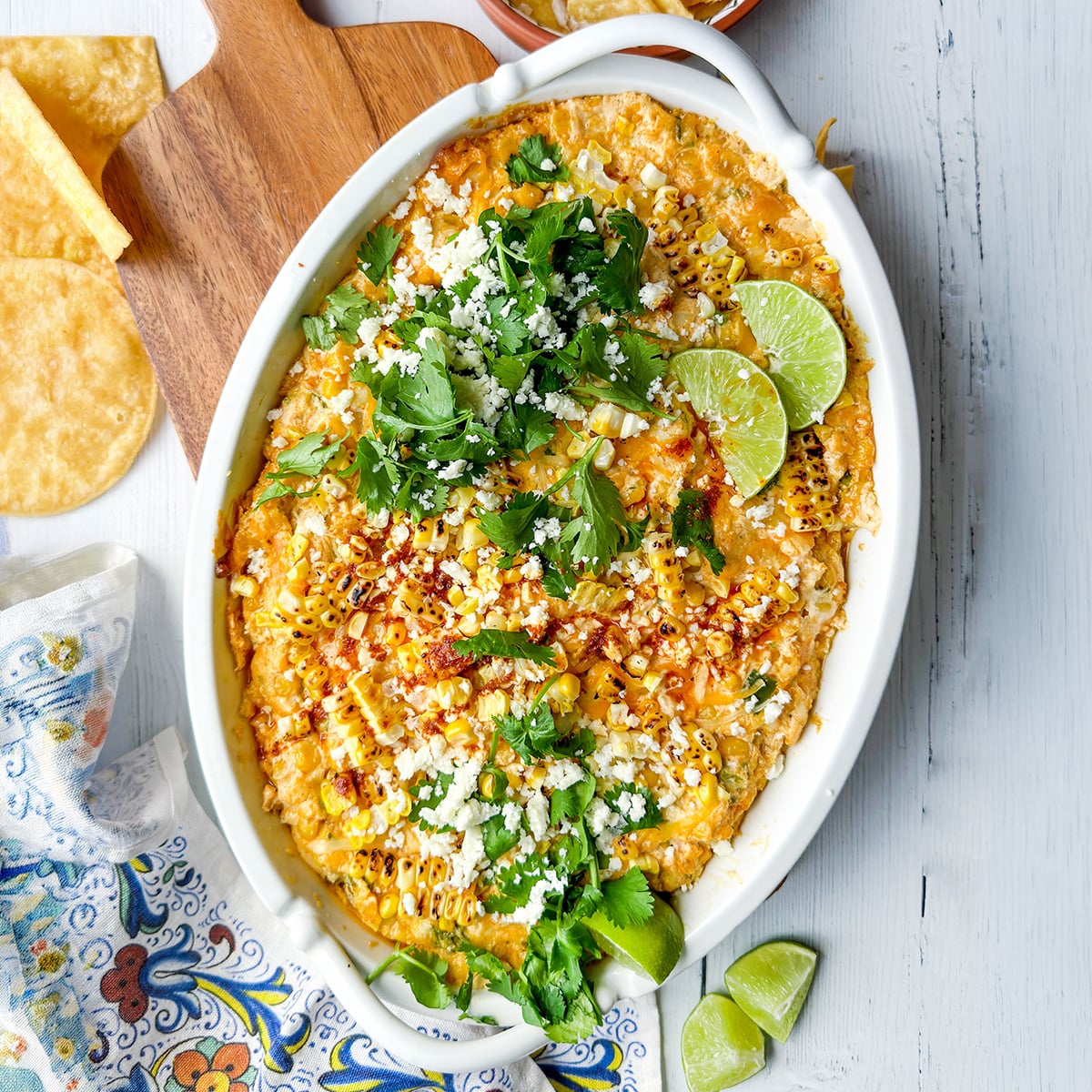 An oval dish of cheesy corn dip—perfect for Game Day Dips—topped with cilantro, crumbled cheese, chili powder, and lime wedges on a white wooden surface.