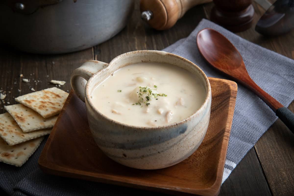 A ceramic mug brimming with creamy fish chowder, garnished with herbs, sits on a wooden plate. Accompanying it are several square crackers and a wooden spoon, all arranged on a gray cloth over the rustic wooden table.