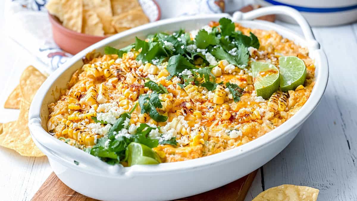 A white casserole dish filled with baked Mexican street corn dip, topped with grilled corn, cilantro, cheese, lime wedges-perfect for serving with tortilla chips at your next Game Day Dips lineup.