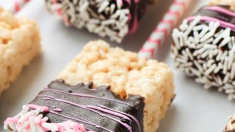 Rectangular Rice Krispies treats on straws, partially dipped in chocolate and decorated with white and pink sprinkles plus a pink icing drizzle, make festive Valentine's Day Treats displayed on a white surface.