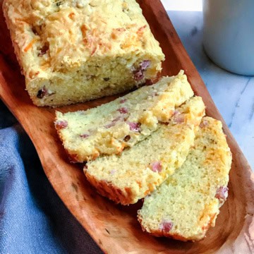 A loaf of savory ham and gruyere quick bread, with visible bits of ham, is partially sliced and displayed on a wooden tray.