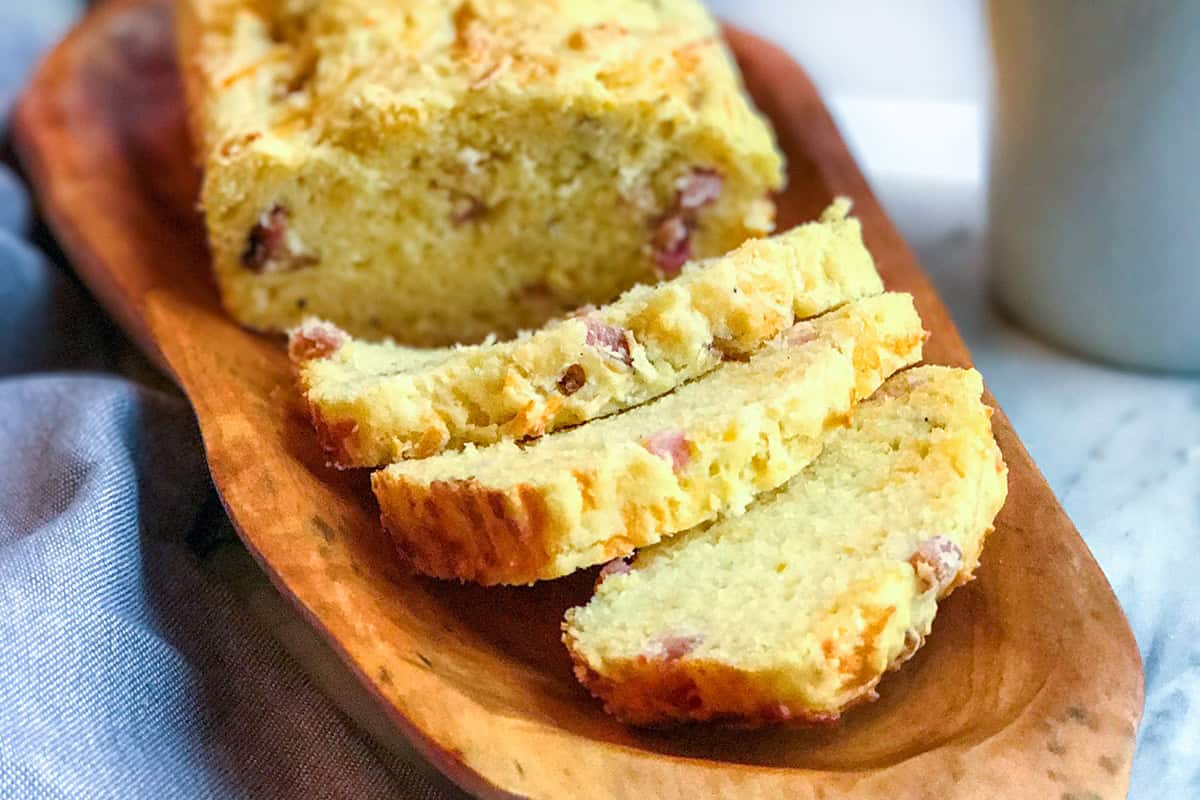 A wooden tray with a loaf of ham and gruyere quick bread, partially sliced. The bread has a light, crumbly texture with visible pieces of ham.