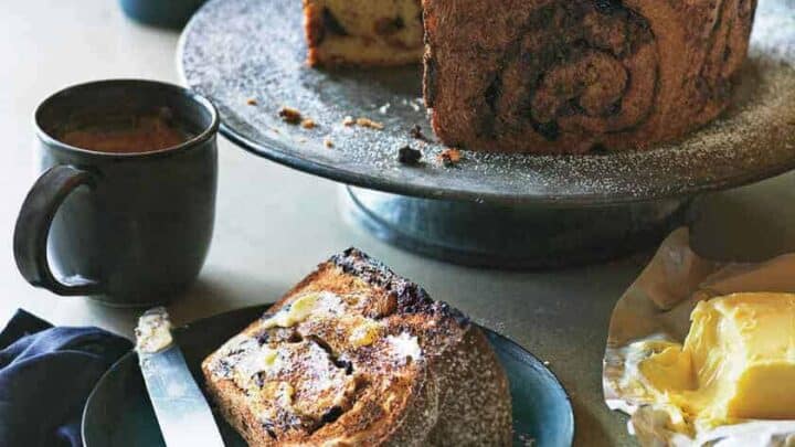 Sliced chocolate babka on a blue plate with a knife, next to a full babka on a raised stand. Two mugs of coffee and a dish with butter complete this cozy Valentine's Day breakfast scene on the marble countertop.
