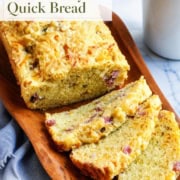 Slices of ham and Gruyere quick bread rest on a wooden tray beside a blue cup and matching linen napkin.