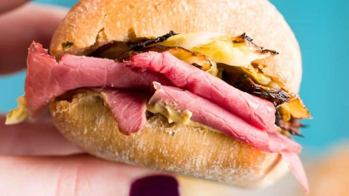 A hand with dark nail polish holds a small sandwich, layered with slices of pink meat and grilled onions between a golden-brown bun. The scene, reminiscent of browsing cabbage recipes, is set against a blurred blue and beige background.