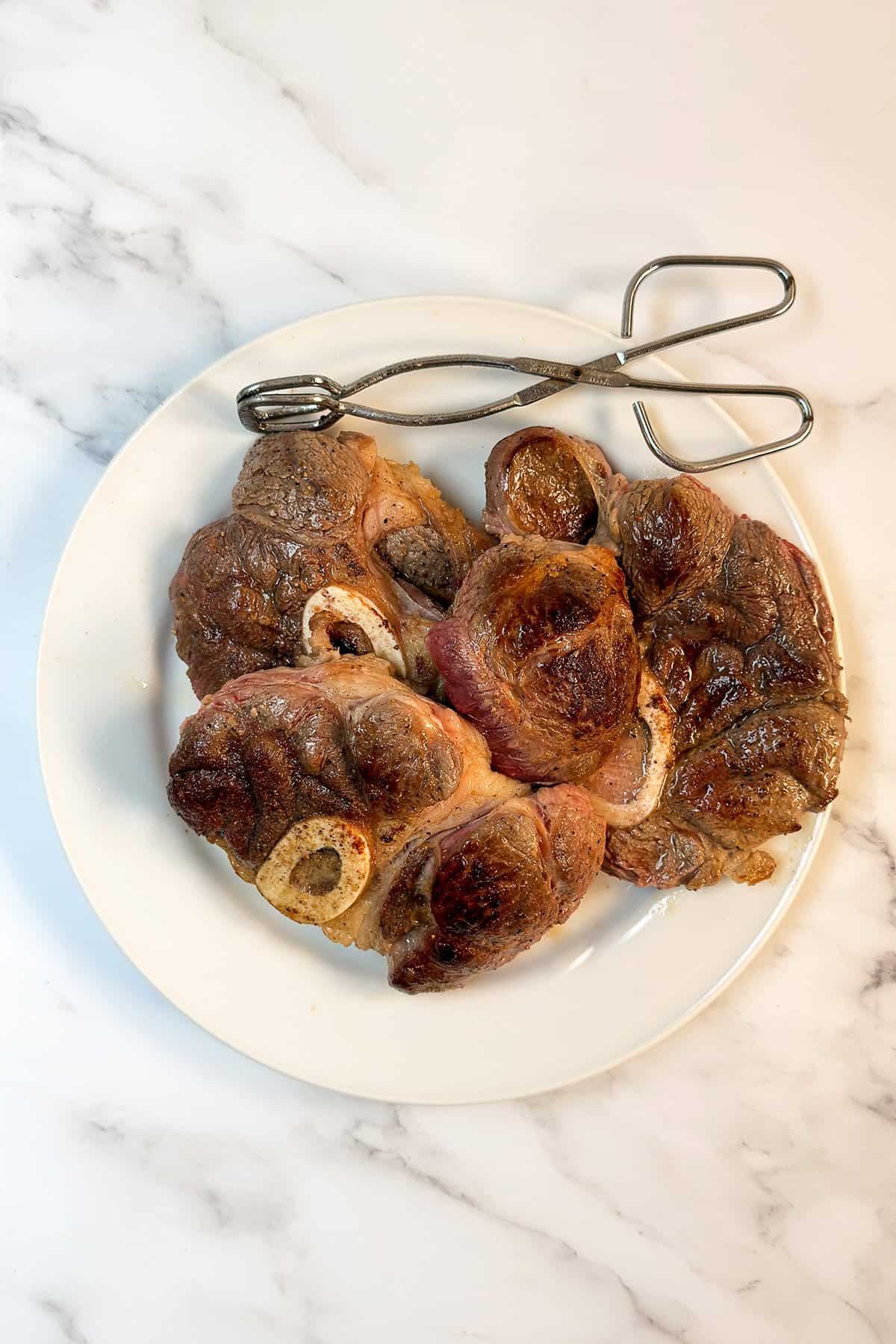 A white plate with several pieces of slow cooker osso buco topped with browned crust, placed on a marble surface, with metal tongs resting on the edge of the plate.