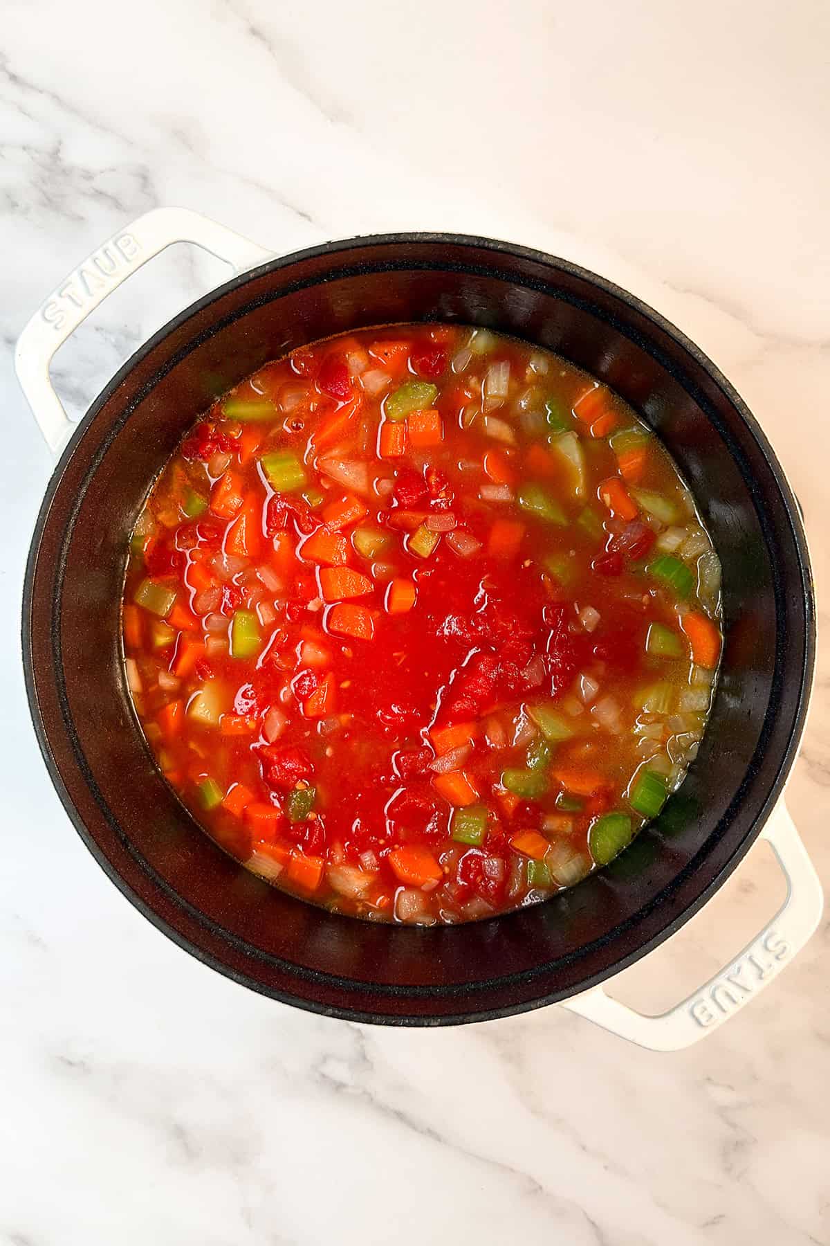 A pot of vegetable soup with carrots, celery, green bell pepper, and diced tomatoes simmers on a white marble surface, reminiscent of the comforting flavors found in slow cooker osso buco.