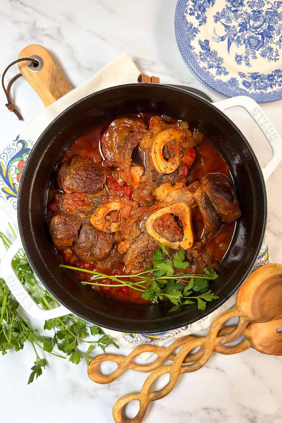 A pot of slow cooker osso buco with beef shanks, bone marrow, and tomato sauce garnished with fresh parsley sits on a marble countertop, surrounded by utensils and plates.