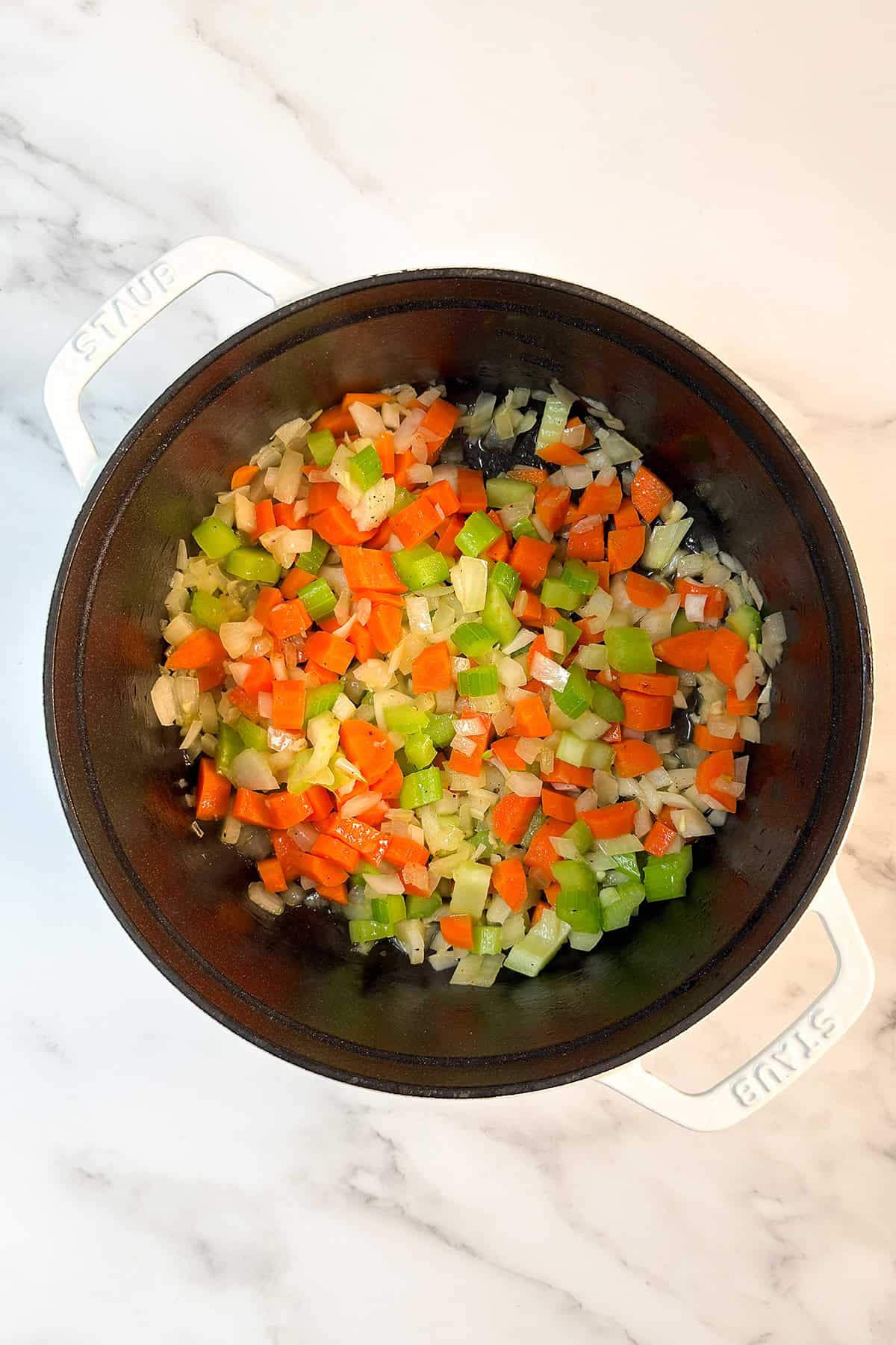 Chopped carrots, celery, and onions are saut&eacute;ing in a white Dutch oven on a marble countertop-ideal for starting a flavorful slow cooker osso buco.