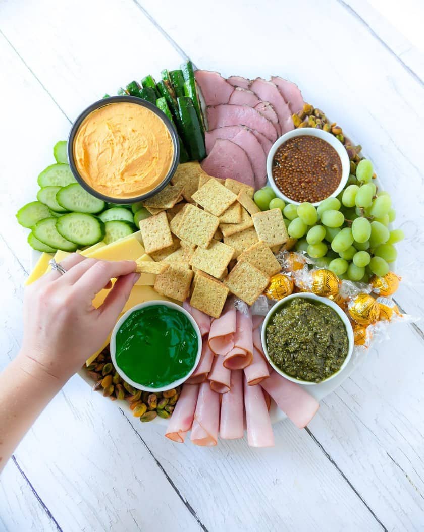 A festive charcuterie board with sliced meats, cheeses, crackers, dips, mustard, grapes, chocolates, cucumbers, and a hand reaching for green jelly-perfect for serving as St Patrick's Day appetizers.