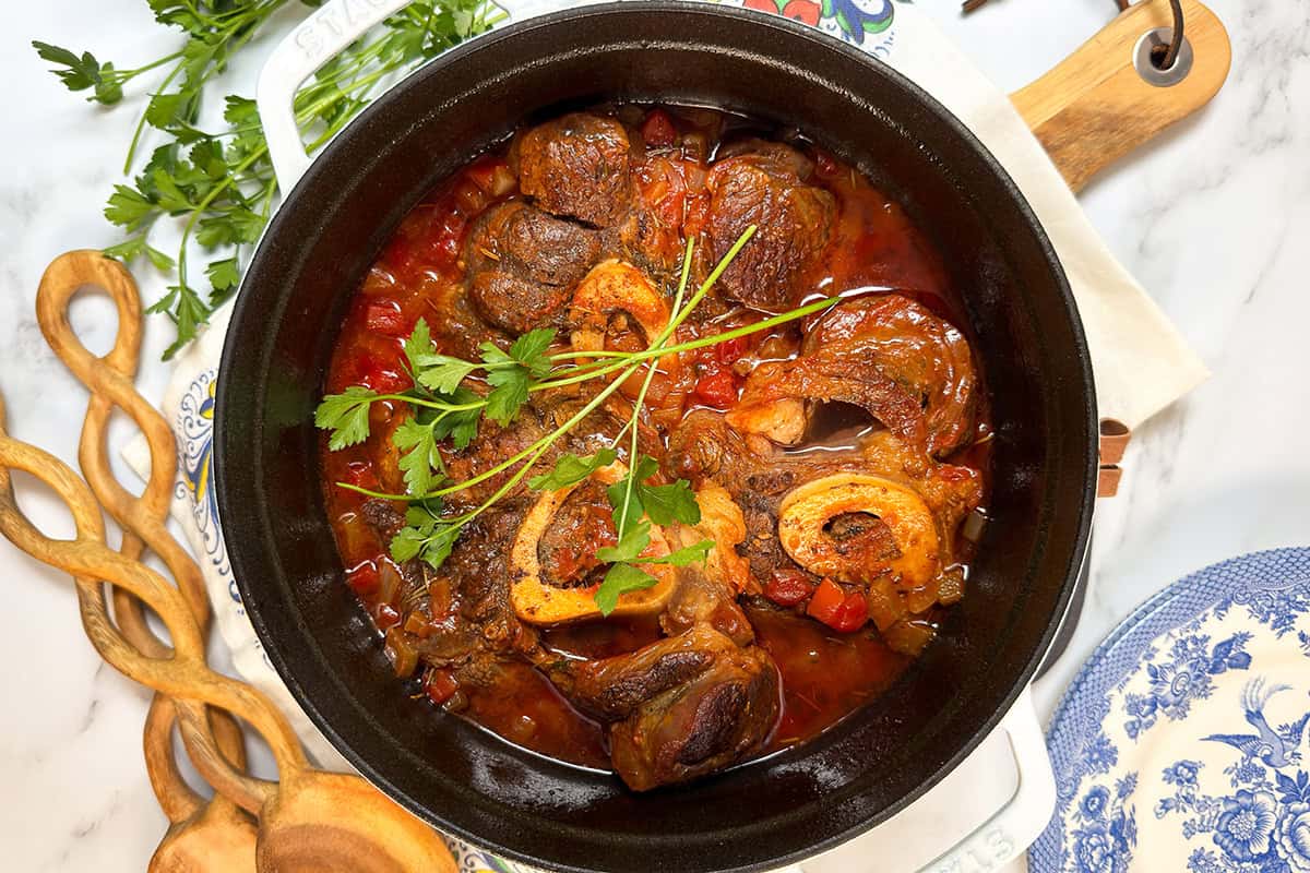 A pot of slow cooker osso buco with beef shanks, bone marrow, and vegetables, garnished with fresh parsley, sits on a marble countertop beside serving utensils and a blue-patterned plate.